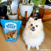 A white dog standing next to a bag of Duck Feet Dog Treats (25 Pack) from Best Bully Sticks.