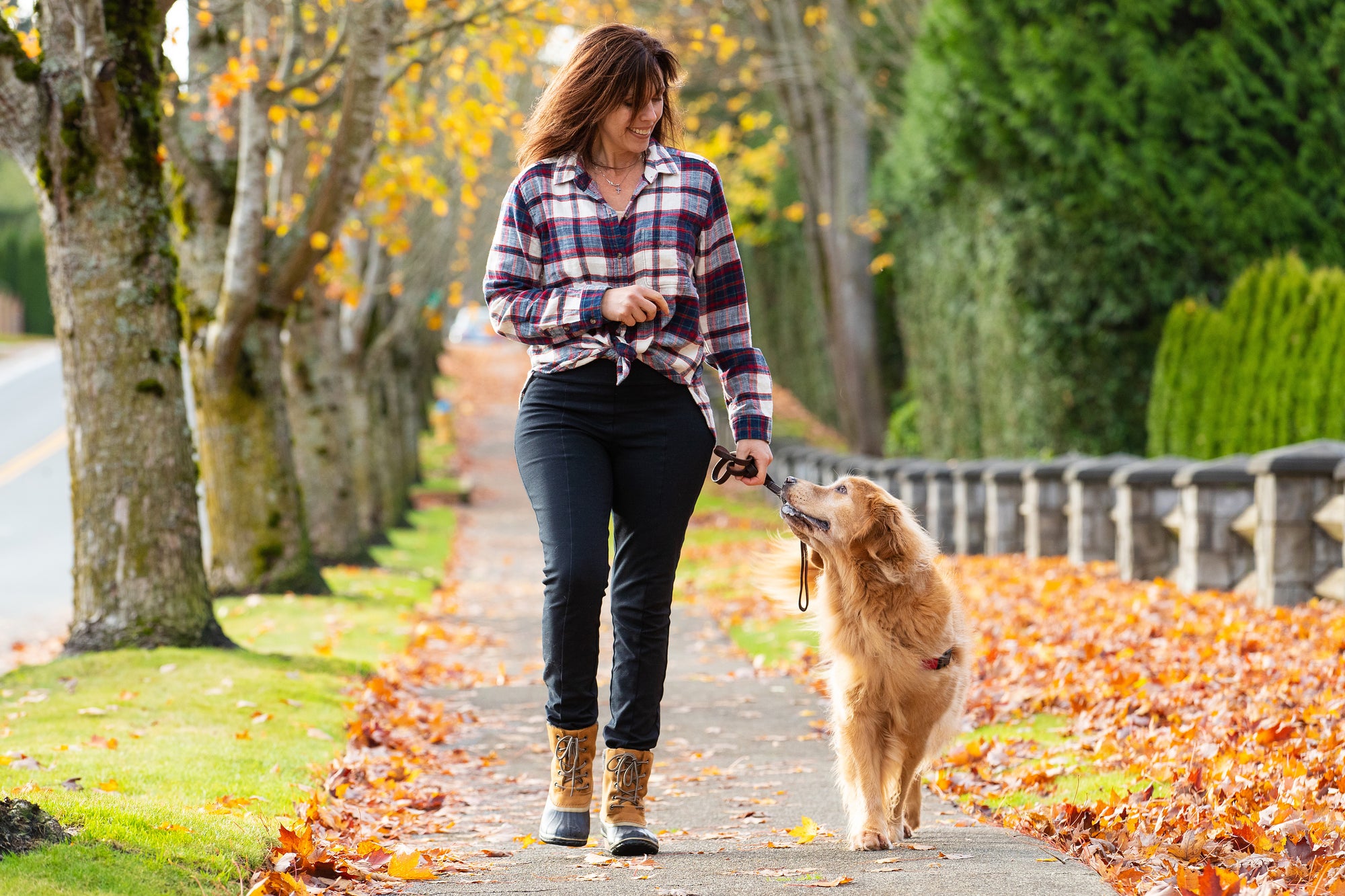 a woman walking her dog on a sidewalk with leaves on the grass